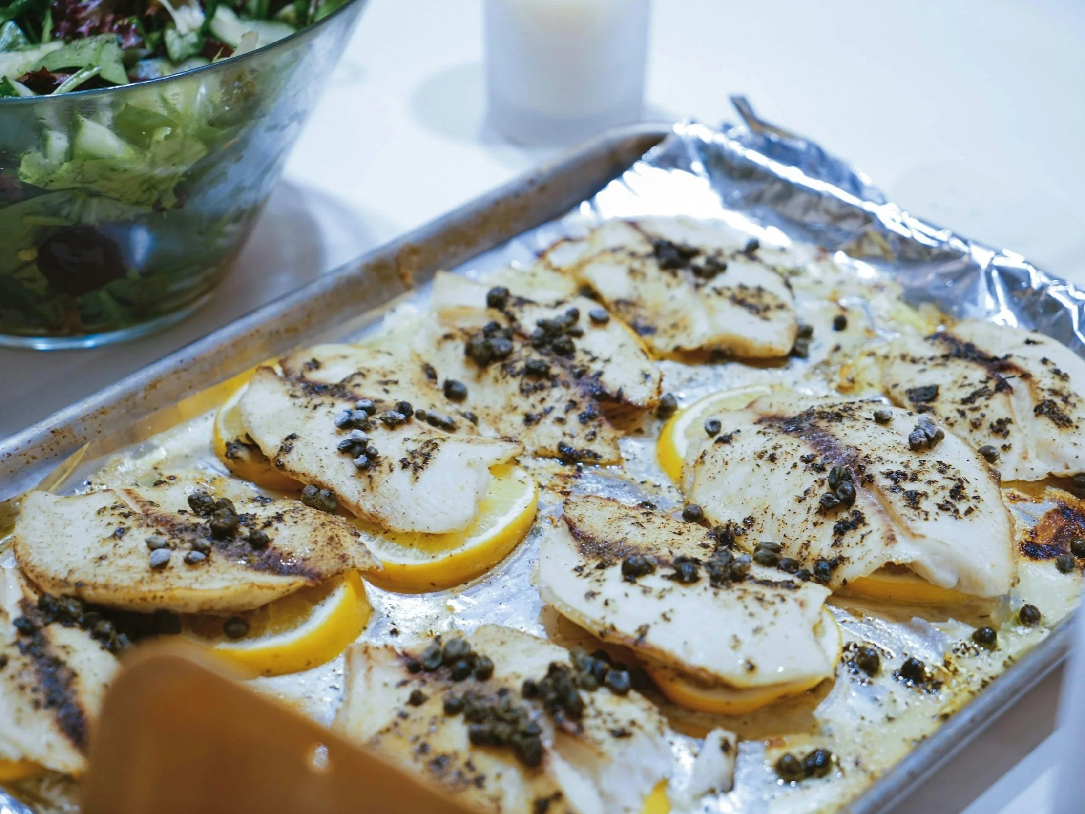 unsplash-image-iJMX64kqJZA Baked fish fillets seasoned with herbs and capers on a tray, served alongside a fresh green salad in a bowl.